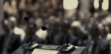 Two podium microphones set up on a stage facing a blurred conference audience